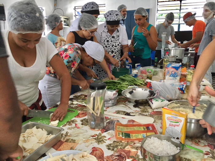 Participante da aula prática, ao redor de uma mesa, com vários legumes e frutas, fazendo receitas gastronômicas